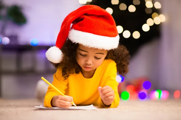 Criança africana escrevendo lista de desejos, deitada no chão perto da árvore de Natal — Fotografia de Stock