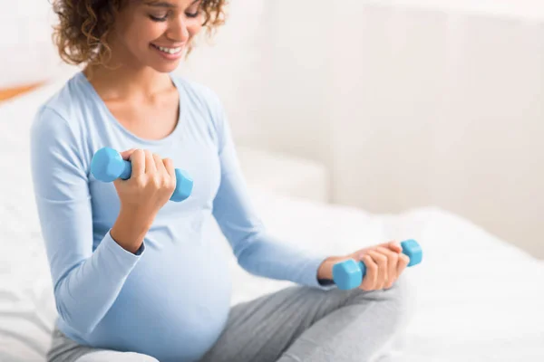 African pregnant lady exercising with dumbbells, sitting on bed — Stock Photo, Image