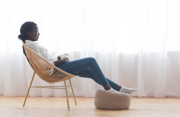 Афроженщина отдыхает в плетеном кресле и пьет кофе у окна — стоковое фото