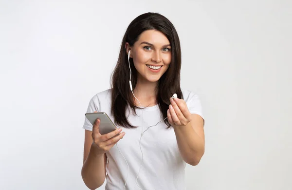Chica sosteniendo Smartphone que ofrece auriculares escuchando música, fondo blanco —  Fotos de Stock