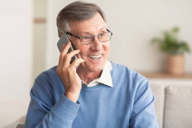 Evde koltukta oturan, cep telefonuyla konuşan yaşlı adam.