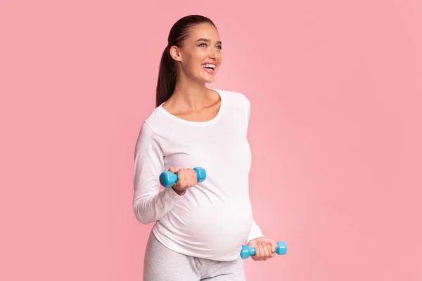 Улыбающаяся беременная женщина упражняется с гантелями, стоящими на розовом фоне — стоковое фото