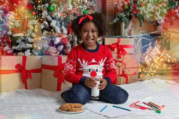 Menina afro bonito beber leite e comer biscoitos perto da árvore de Natal — Fotografia de Stock