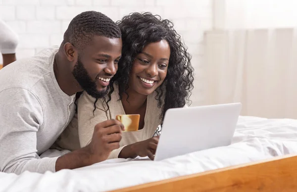 Feliz pareja afroamericana de compras en línea, utilizando el ordenador portátil y la tarjeta de crédito en la cama, espacio vacío —  Fotos de Stock