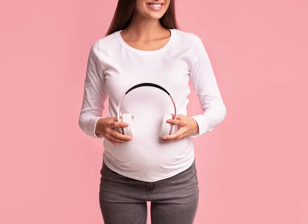 Nerozpoznatelná očekávaná dívka drží sluchátka v blízkosti břicha, růžové pozadí, oříznuté — Stock fotografie