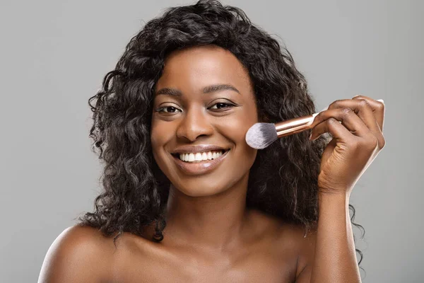 Menina preta atraente usando ferramenta de escova, aplicando blush no rosto — Fotografia de Stock