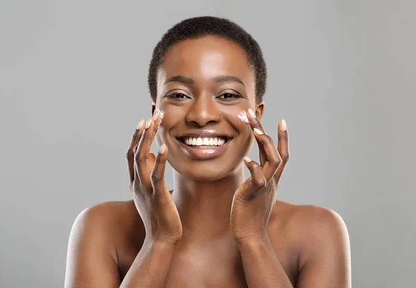 Retrato de mulher negra feliz aplicando creme hidratante no rosto — Fotografia de Stock