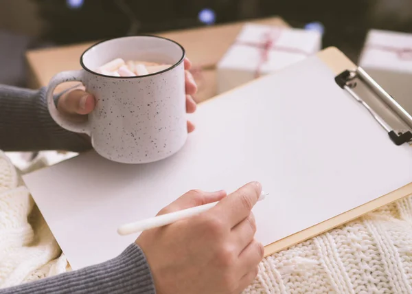 Woman drinking hot chocolate and writing greetings on white clipboard — Stock Photo, Image