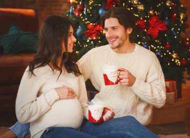 Evli çift kakao içiyor ve Noel ağacının yanında konuşuyor.