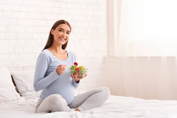 Wanita hamil memegang mangkuk dengan salad duduk di tempat tidur di dalam ruangan — Stok Foto