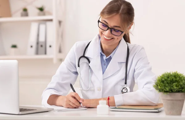 Lächelnde Ärztin verschreibt Medikamentenbehandlung sitzend am Arbeitsplatz — Stockfoto