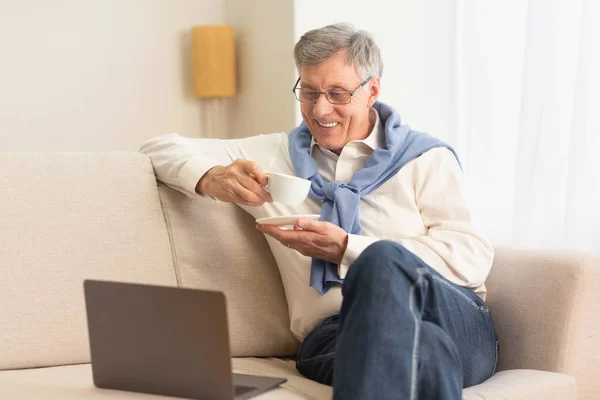 Ηλικιωμένος άνθρωπος χρησιμοποιώντας το φορητό υπολογιστή έχοντας καφέ κάθεται στον καναπέ — Φωτογραφία Αρχείου