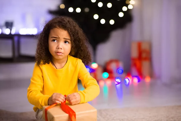 Triste menina afro tentando abrir caixa de presente na véspera de Natal — Fotografia de Stock