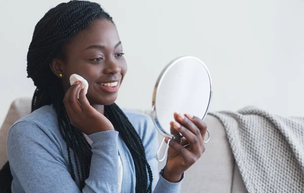 Mujer africana mirando en el espejo y la cara de limpieza con almohadilla de algodón — Foto de Stock