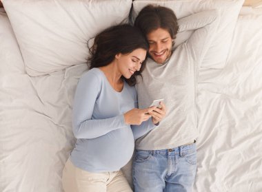 Hamile kadın kocasıyla yatakta dinlenirken akıllı telefon kullanıyor.
