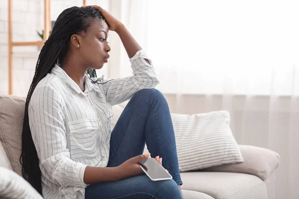 Afro chica molesta sentada en el sofá con el teléfono celular, pensando en algo — Foto de Stock