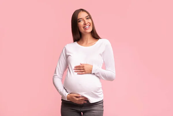 Veselý těhotná dáma dotýkající se břicho stojící na růžovém pozadí — Stock fotografie