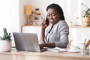Afro iş kadını cep telefonuyla konuşuyor ve ofiste dizüstü bilgisayar kullanıyor.