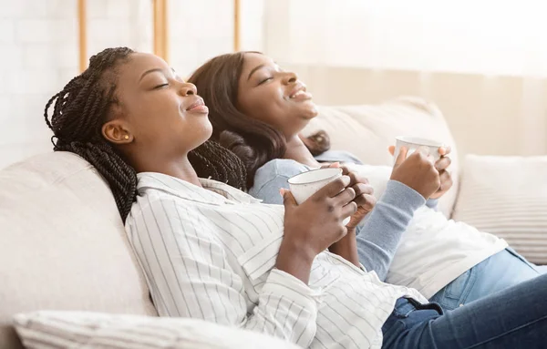 Two african american girlfriends resting on sofa with coffee — Stock Photo, Image