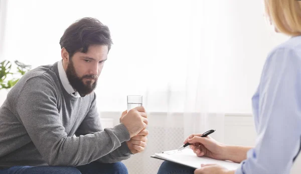 Upset man calming down with glass of water at psychotherapy session