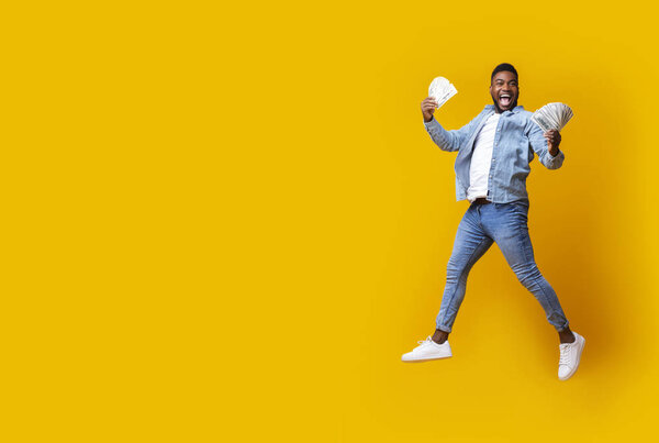 Big Win. Joyful Afro Guy Holding Cash, Jumping In Air And Screaming with Excitement Over Yellow Studio Background With Copy Space