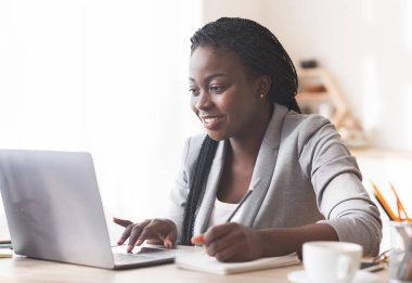 Gülümseyen siyah iş kadını laptopta çalışıyor ve ofiste notlar alıyor..