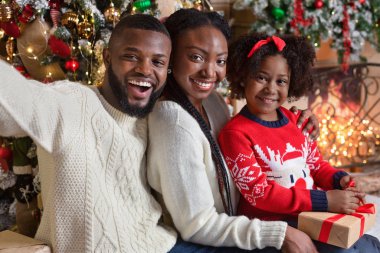 Siyah ebeveynler kızlarıyla Noel ağacının yanında selfie çekiyorlar.