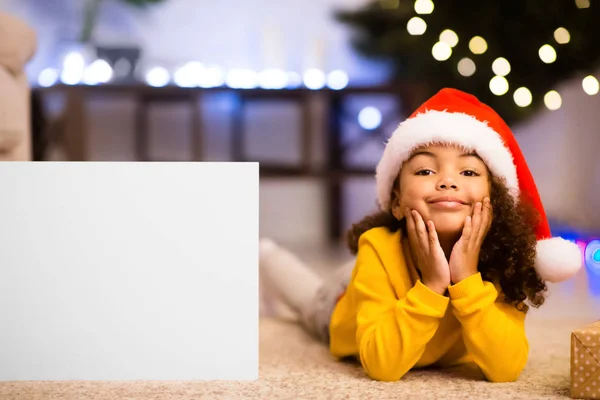 Чарівна маленька дівчинка в капелюсі Санта лежить поруч з порожнім плакатом — стокове фото
