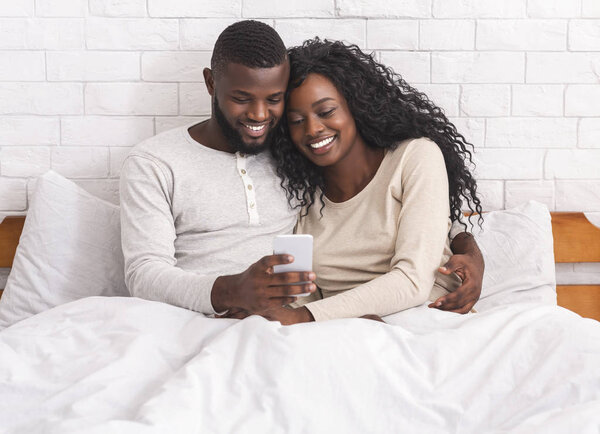Smiling black couple relaxing in bed with smartphone