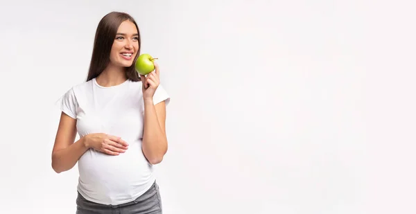 妊娠中の女の子が白いスタジオの背景に立ってアップルを食べる,パノラマ — ストック写真
