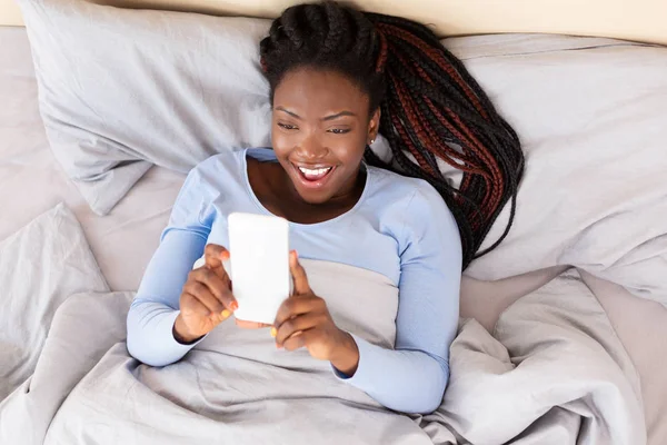 Chica feliz usando teléfono sosteniendo las manos por encima de acostado en la cama —  Fotos de Stock