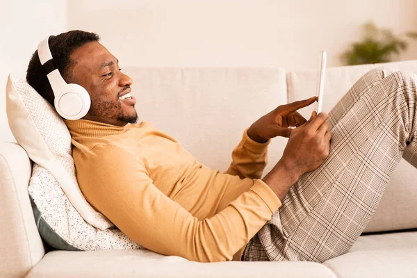 Afro Man fülhallgató használata Tablet feküdt a kanapén — Stock Fotó