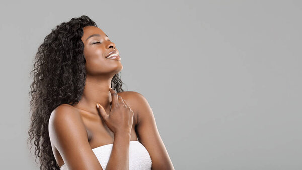 Body care concept. Pleased beautiful black woman enjoying touching her soft skin, standing wrapped in towel after spa treatment over gray background