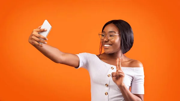 Afro ragazza making selfie su smartphone gesturing v-sign, sfondo arancione — Foto Stock