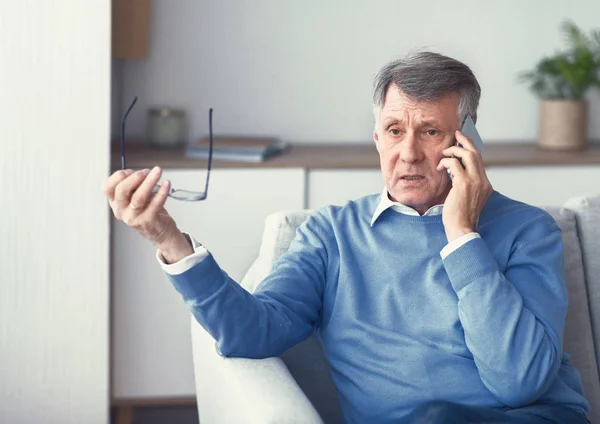 Старший человек, имеющий неприятный телефонный разговор сидит на диване в помещении — стоковое фото