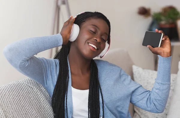 Веселая черная девушка в наушниках слушает музыку на смартфоне дома — стоковое фото