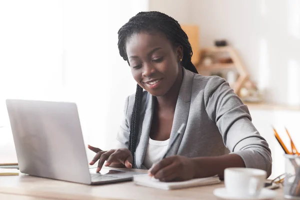 Donna d'affari nera che lavora in ufficio, digitando sul computer portatile e prendendo appunti — Foto Stock