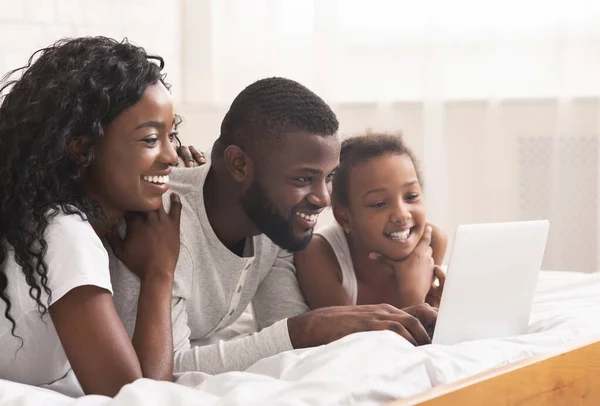 Alegre familia negra de tres usando el ordenador portátil en la cama juntos —  Fotos de Stock