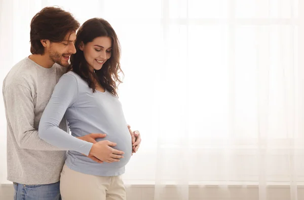 Feliz esperando casal abraçando enquanto está perto da janela em casa — Fotografia de Stock