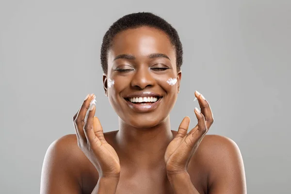 Mulher afro bonita aplicando creme em suas bochechas e sinceramente sorrindo — Fotografia de Stock
