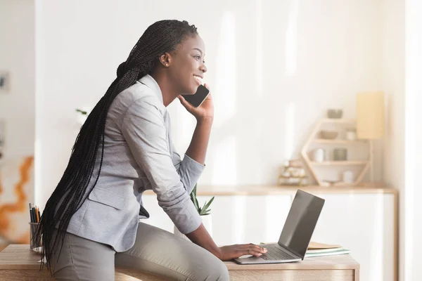 Νεαρή μαύρη επιχειρηματίας που μιλάει στο κινητό και χρησιμοποιεί φορητό υπολογιστή στο γραφείο — Φωτογραφία Αρχείου