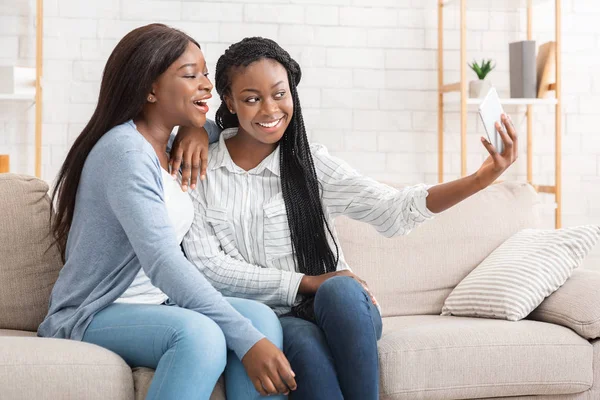 Cheerful black girls taking selfie while sitting on couch at home — ストック写真