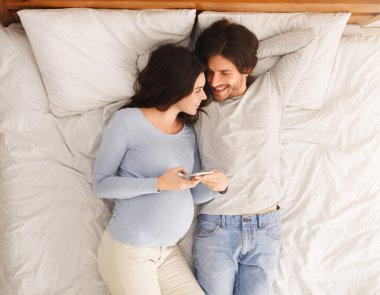 Kocasıyla yatakta yatarken akıllı telefonu tutan hamile bir kadın.