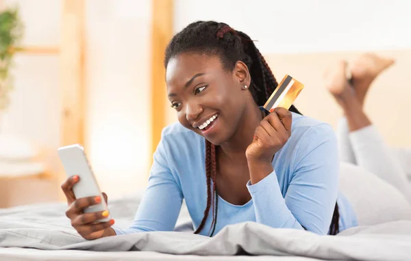 Счастливая чернокожая женщина, пользующаяся телефоном при покупке онлайн, лежа в кровати — стоковое фото
