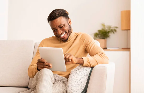 Siyah adam tablet kullanıyor, gülümsüyor evde koltukta oturuyor. — Stok fotoğraf