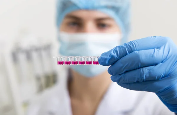 Розмита лабораторна асистентка в рукавицях дивиться на рожеву рідину в пробірках — стокове фото