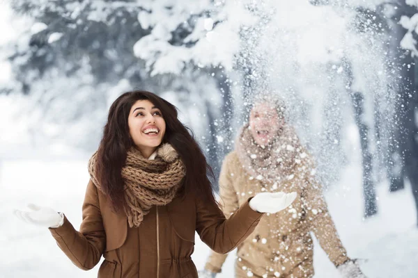 Mujer alegre lanzando nieve caminando con su novio en el bosque nevado —  Fotos de Stock