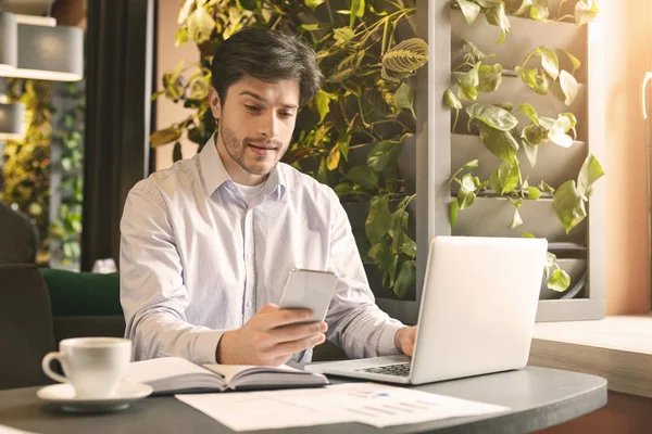 Millennial man chatten op mobiele telefoon, werken aan laptop in cafe — Stockfoto