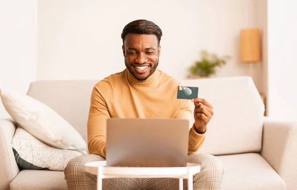 Chico feliz sosteniendo tarjeta de crédito usando el ordenador portátil sentado en casa —  Fotos de Stock
