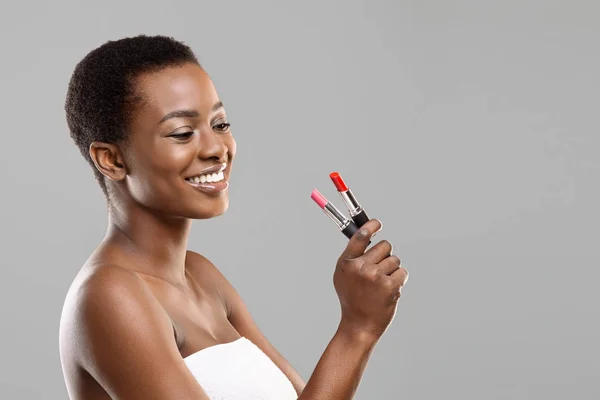 Bonita chica negra sosteniendo y eligiendo entre lápiz labial rojo y rosa — Foto de Stock